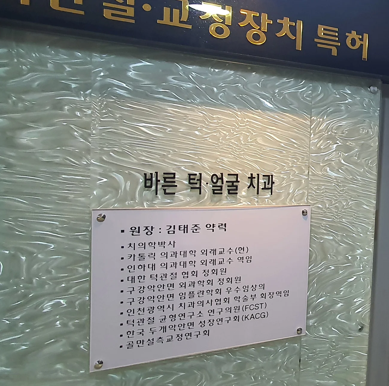 바른턱얼굴치과의원 입구에 게시된 김태준 대표원장 약력 명판 — 치의학박사, 카톨릭 의과대학 외래교수(현), 인하대 의과대학 외래교수 역임, 대한 턱관절 협회 정회원, 구강악안면 외과학회 정회원, 구강악안면 임플란트학회 우수임상의, 인천광역시 치과의사협회 학술부 회장역임, 턱관절 균형연구소 연구의원(FCST), 한국 두개악안면 성장연구회(KACG), 골만설측교정연구회